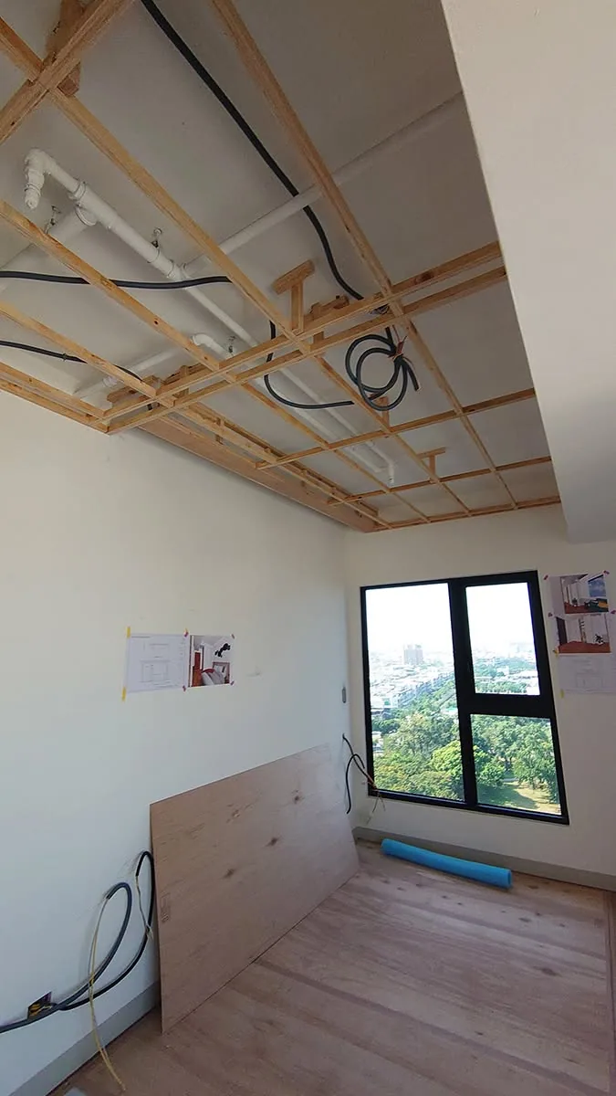 臥室天花板角材骨架間可見白色排水管與黑色電線,牆面張貼設計圖