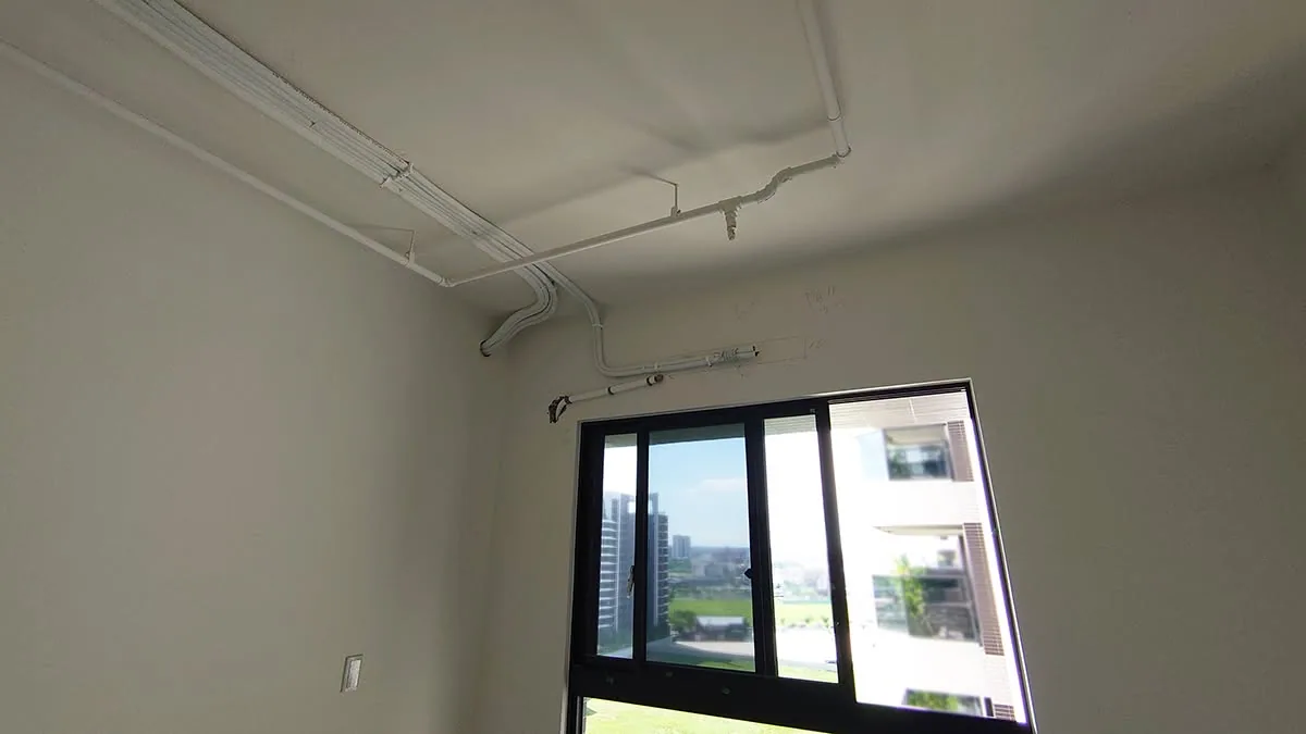 空房間天花與牆面走明管,白色配管沿樑邊轉折至窗上方,室內僅有鋁框推拉窗