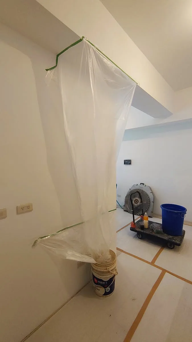 施工現場以塑膠布與膠帶做牆面保護,地面鋪設保護板,旁邊放置油漆桶與送風機