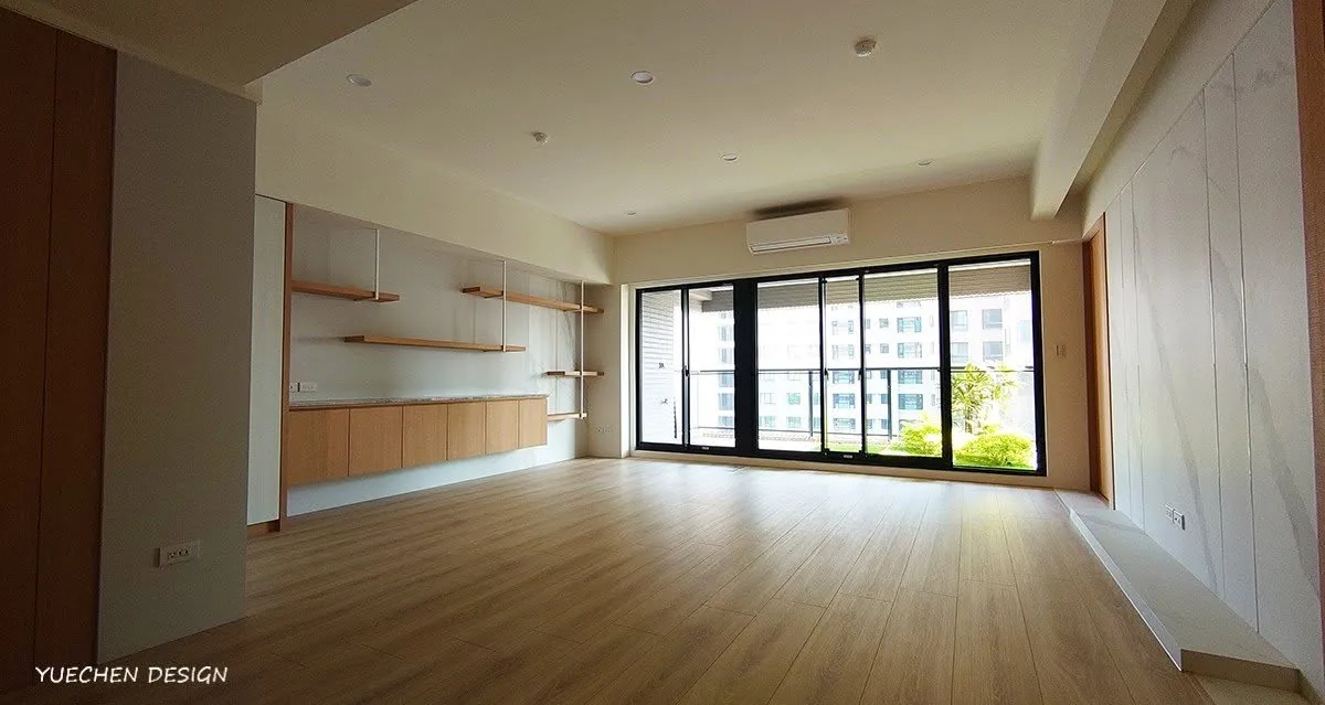 空屋客廳鋪淺色木地板，牆面設層板與矮櫃，落地窗外可見社區綠意